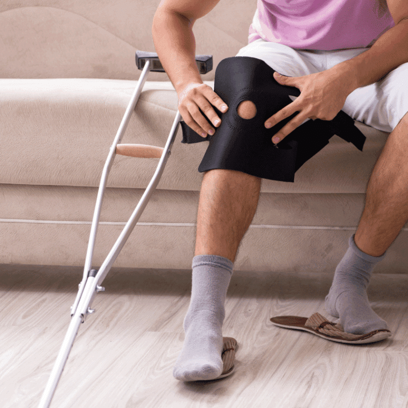 Man sitting on a couch adjusting a black knee brace with crutches beside him